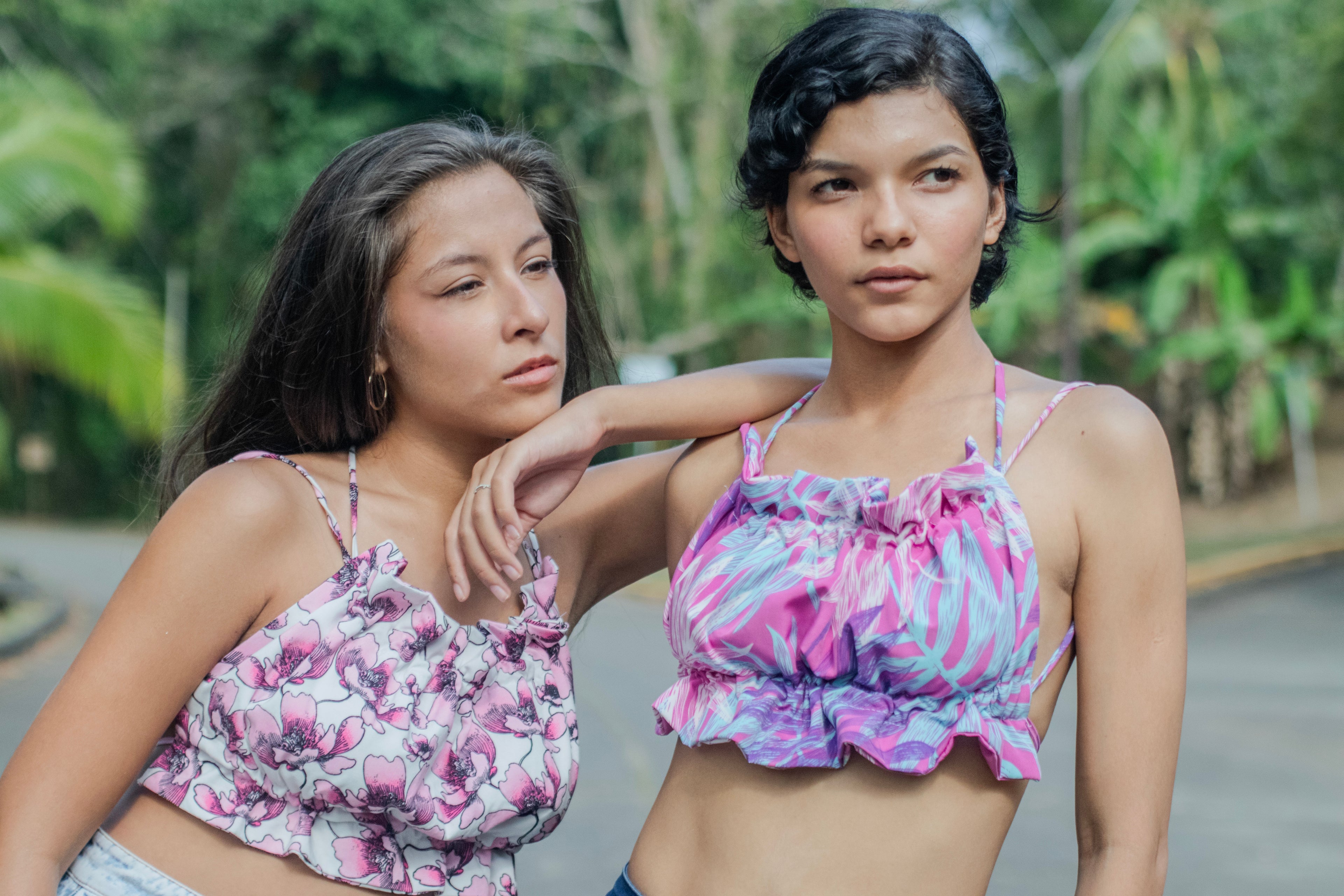 Dos modelos posando al aire libre, usando blusas de tirantes con detalles fruncidos en telas recicladas, con un fondo natural de árboles y vegetación. La modelo a la izquierda viste una blusa de estampado floral en tonos rosados y la modelo a la derecha lleva una blusa con un diseño abstracto en tonos morados y azules. Ambas transmiten una sensación relajada y fresca, destacando la moda sostenible y creativa.
