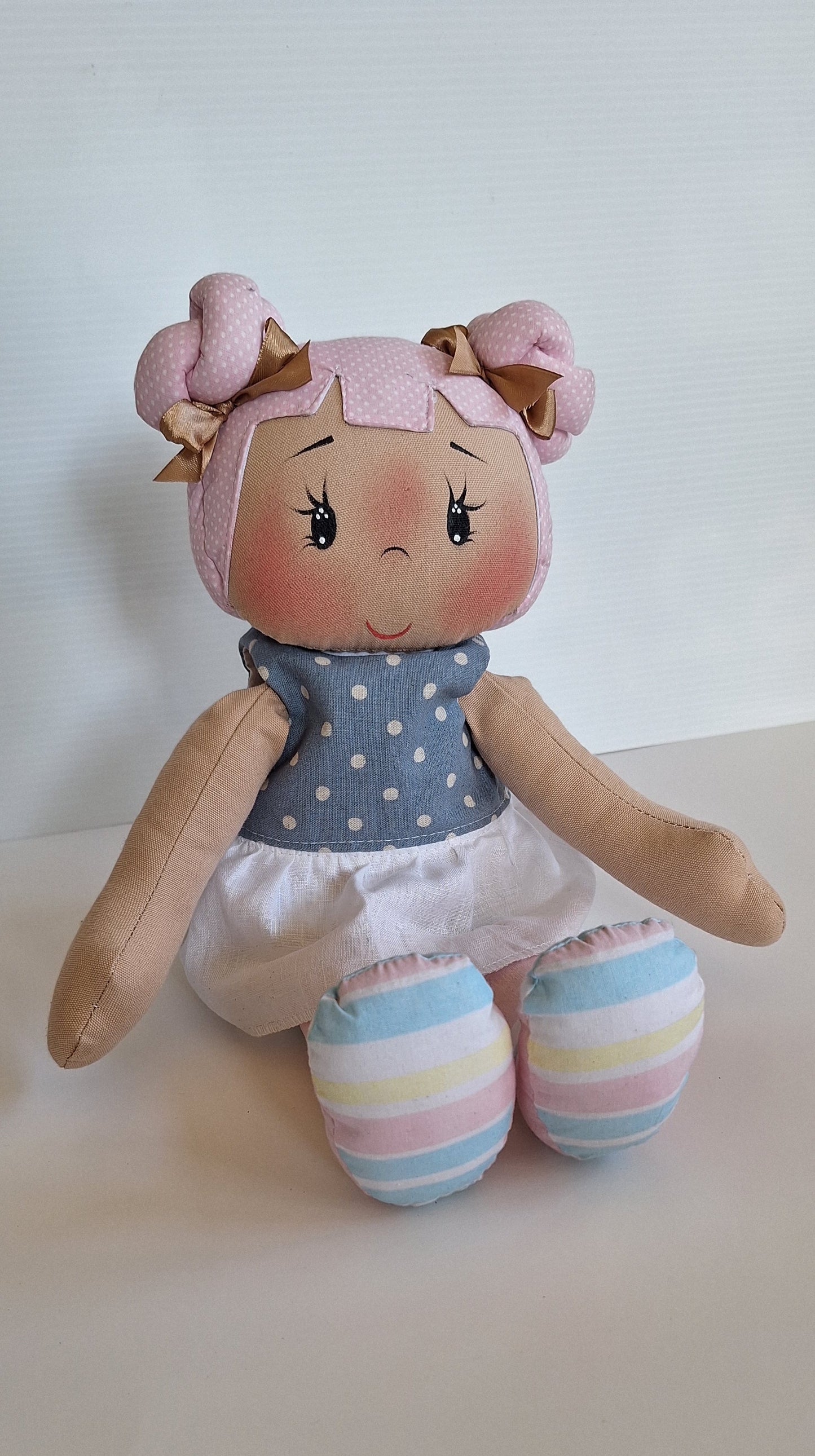 Muñeca de tela artesanal sentada, con cabello rosa trenzado adornado con moños dorados, blusa de lunares blancos, falda blanca y piernas de rayas pastel, sobre fondo blanco.